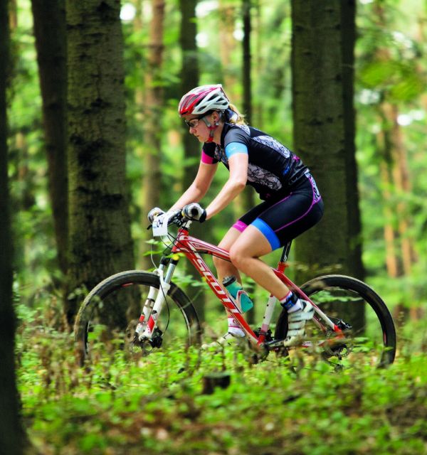 Kobieta w profesjonalnym stroju kolarskim pokonuje na rowerze leśną trasę terenową. Do kierownicy ma przypięty numer startowy. Wokół drzewa.