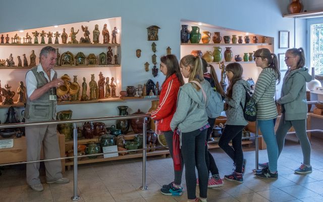Grupa dziewczynek w wieku szkolnym zwiedza wystawę ośrodka tradycji garncarstwa w Chałupkach. Towarzyszy im przewodnik, który opowiada o jednym z eksponatów.